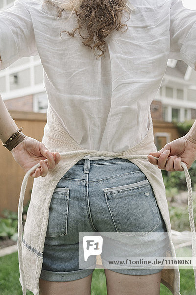 A woman in denim shorts tying a work apron  back view.