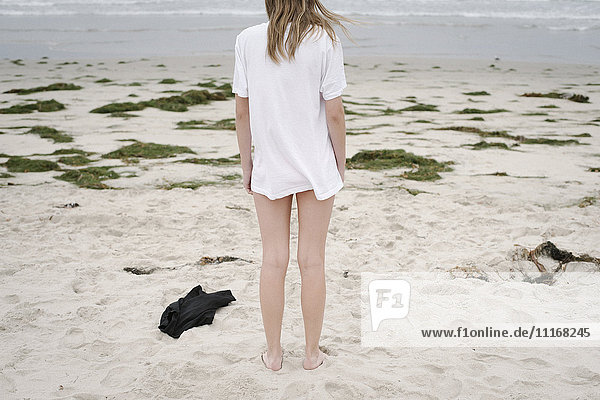 Rückansicht eines Mädchens mit blonden Haaren in einem weißen T-Shirt  das an einem Sandstrand steht.