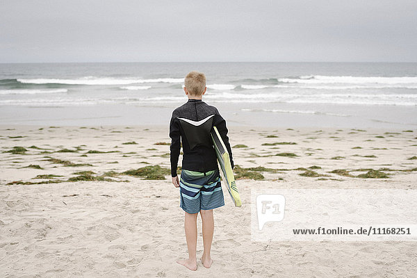 Blond boy standing on a sandy beach  holding a bodyboard.