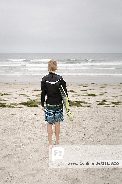 Blond boy standing on a sandy beach  holding a bodyboard.