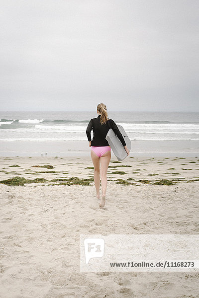 Rückansicht eines Mädchens  das ein Bodyboard trägt und in den Ozean geht.