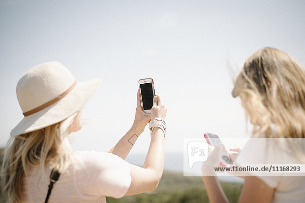 Frau und junges Mädchen mit langen blonden Haaren stehen im Freien  halten ein Mobiltelefon hoch und machen ein Foto.