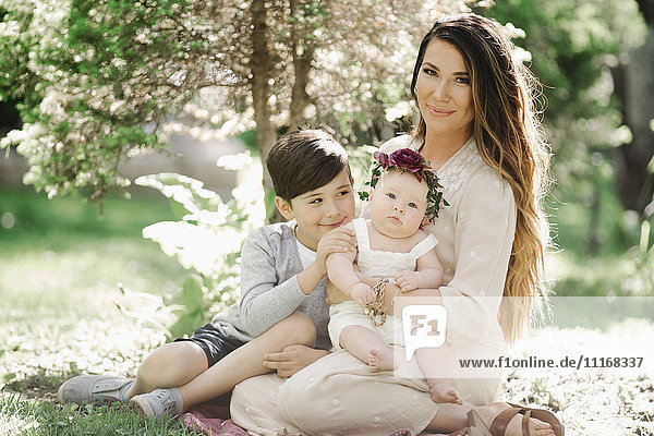 Porträt einer lächelnden Mutter  eines Jungen und eines kleinen Mädchens mit einem Blumenkranz auf dem Kopf  die in einem Garten sitzen.