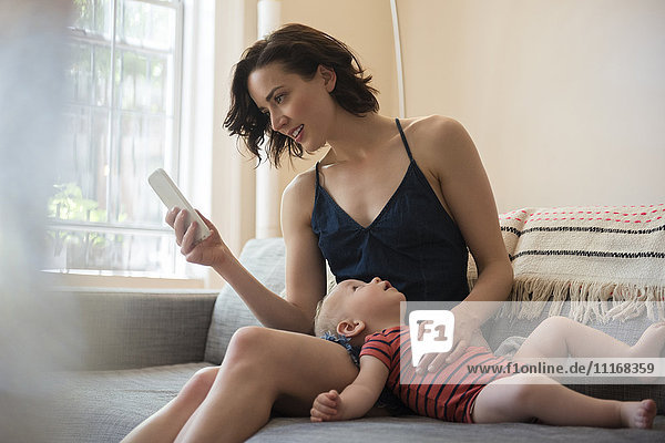 Caucasian mother sitting on sofa with baby son texting on cell phone