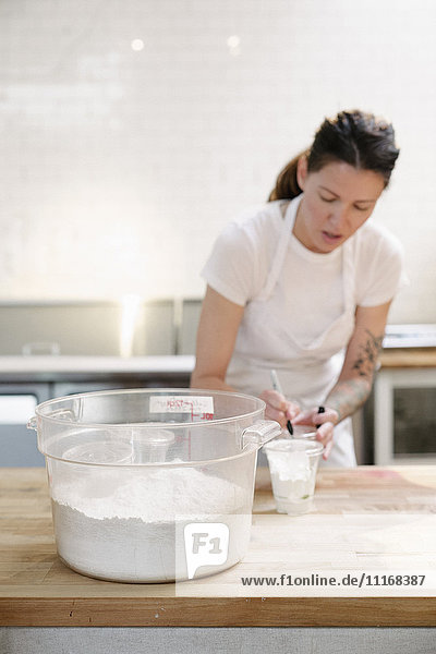 Frau mit weißer Schürze  die an einem Arbeitstresen in einer Bäckerei steht und Mehl misst.
