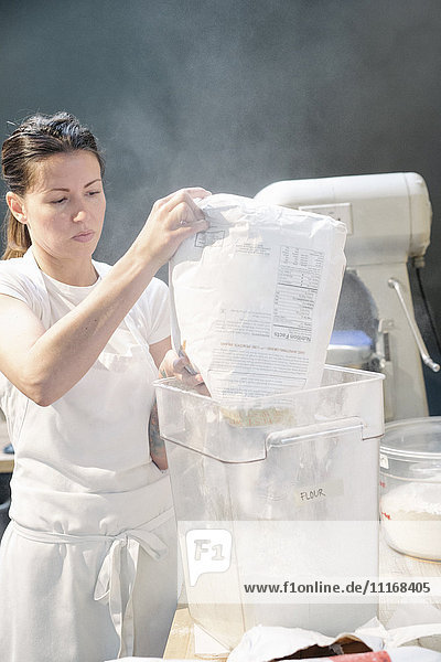 Frau mit weißer Schürze steht an einem Arbeitstresen in einer Bäckerei und schüttet Mehl in einen Behälter.