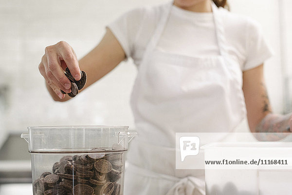 Nahaufnahme einer Frau mit einer weißen Schürze  die an einem Arbeitstresen in einer Bäckerei steht und Schokolade in der Hand hält.
