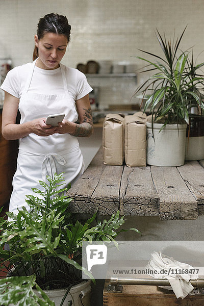 Frau mit weißer Schürze  die in einer Küche steht und ein Mobiltelefon benutzt.