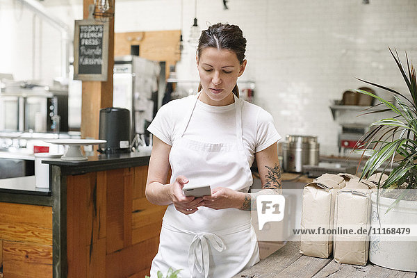 Woman wearing a white apron standing in a kitchen  using a mobile phone.