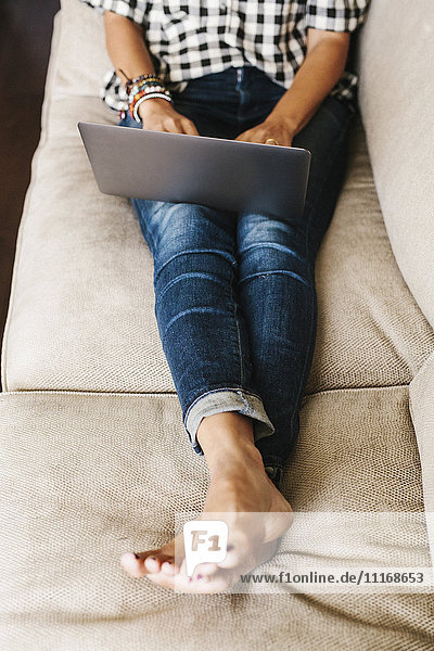 Frau in Jeans  die auf einem Sofa liegt und einen Laptop benutzt.