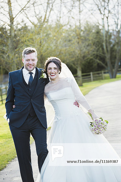 A bride and groom on their wedding day walking arm in arm down a path in the sunshine laughing.