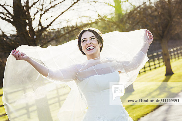 Eine Braut in ihrem Brautkleid,  die an ihrem Hochzeitstag lacht und den Schleier hinter sich heraushält.