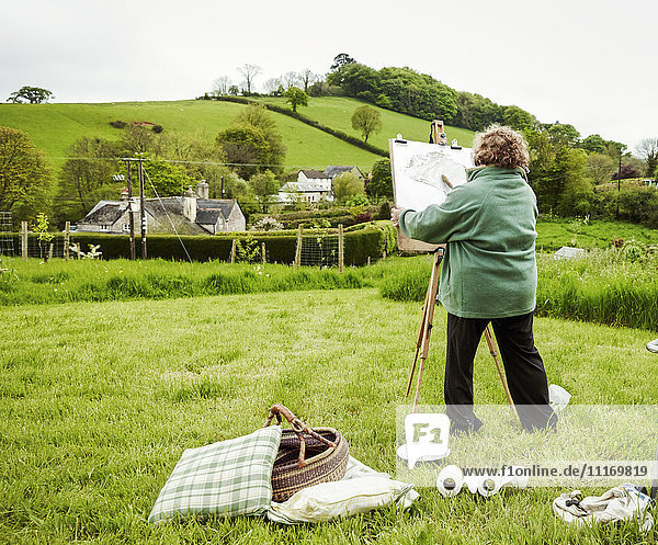 Eine Künstlerin steht im Freien an einer Staffelei und malt eine ländliche Szene.