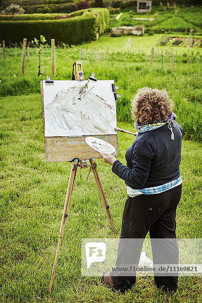 Eine Künstlerin steht im Freien an einer Staffelei und trägt mit einem Pinsel Farbe auf Papier auf.