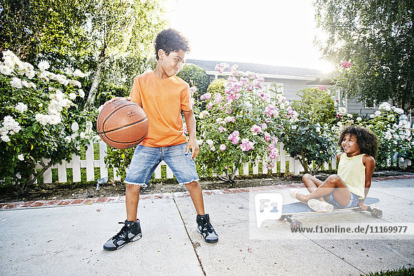 Gemischtrassiges Mädchen auf Skateboard  das seinem Bruder beim Basketballspielen zusieht