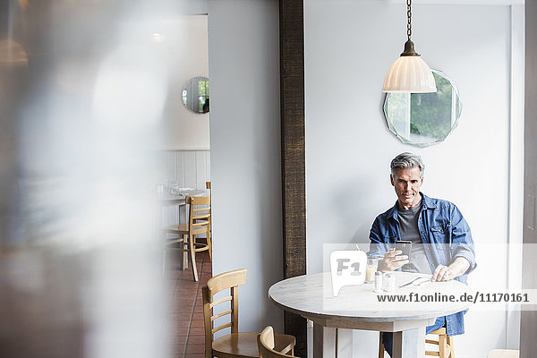 Ein Mann sitzt an einem Coffeeshop-Tisch und schaut auf sein Smartphone. Unscharfer Vordergrund.