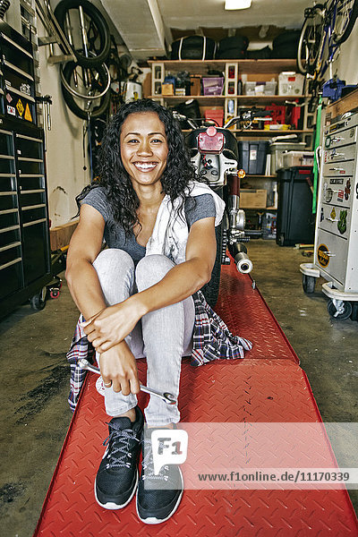Lächelnde gemischtrassige Frau sitzt auf einem Reparaturstand in einer Garage