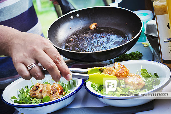 Woman standing at a camping stove  preparing two plates with fried scallops and fresh herbs.