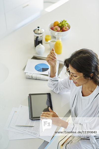 Frau im Bademantel bei der Arbeit am digitalen Tablet am Küchentisch