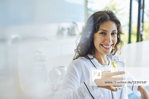 Porträt einer lächelnden Frau im Bademantel  die in der Küche Wasser trinkt