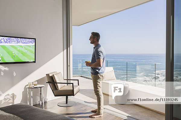 Man watching soccer on TV at sunny luxury patio doorway with ocean view