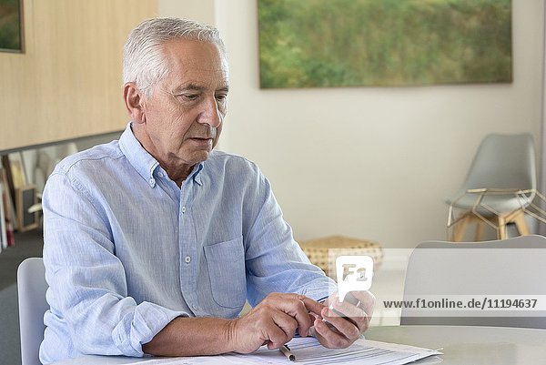 Senior man using a phone while doing paperwork at home