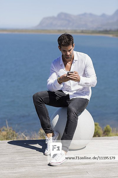 Mittlerer Erwachsener Mann  der auf einer Steinkugel sitzt und ein Smartphone benutzt.