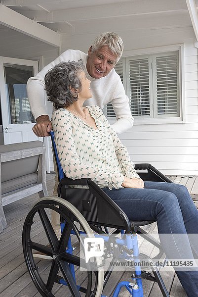 Älterer Mann im Gespräch mit seiner Frau im Rollstuhl auf der Veranda