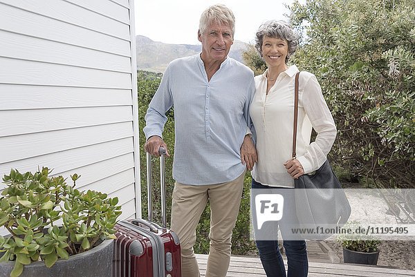Happy senior couple standing with suitcase outside of house