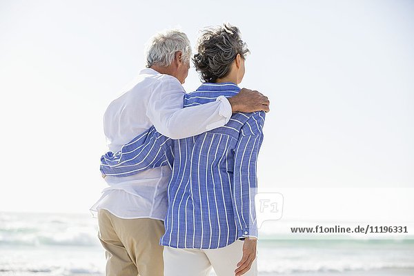 Rückansicht eines Paares  das mit einem Arm um den anderen am Strand steht.