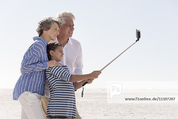 Glückliche Großeltern mit Enkelin  die zusammen am Strand Selfie nehmen.