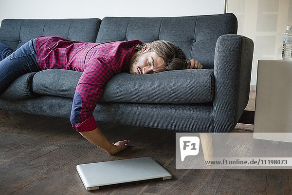 Junger Mann schläft auf Sofa mit Laptop