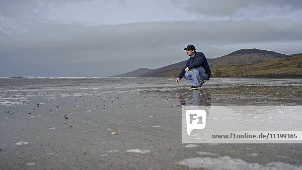 Young Adult Man Crouched on Beach as Waves Roll in
