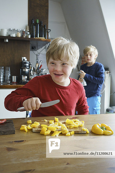 Glücklicher behinderter Junge hackt gelbe Paprika mit Bruder im Hintergrund in der Küche.