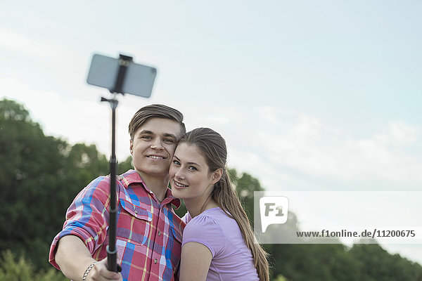Fröhliches junges Paar nimmt Selfie durch Einbeinstativ im Park gegen den Himmel