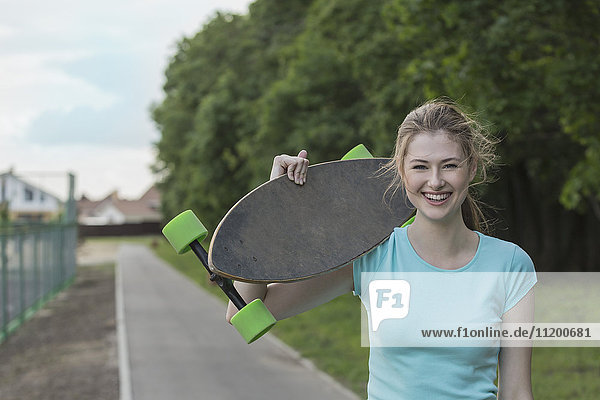 Porträt einer fröhlichen Frau mit einem Skateboard auf einem Fußweg im Park.