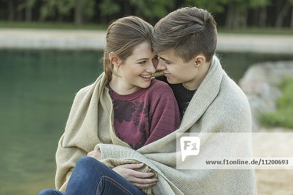 Smiling young loving couple wrapped in blanket resting by lake