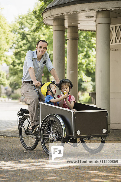 Lächelnder Vater fährt Fahrrad  während Jungen auf der Straße im Wagen sitzen.