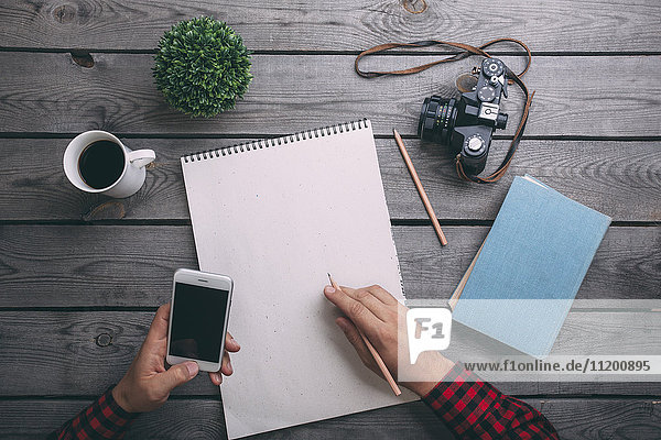 Beschnittenes Bild eines Mannes  der ein Smartphone hält  während er auf Papier schreibt.