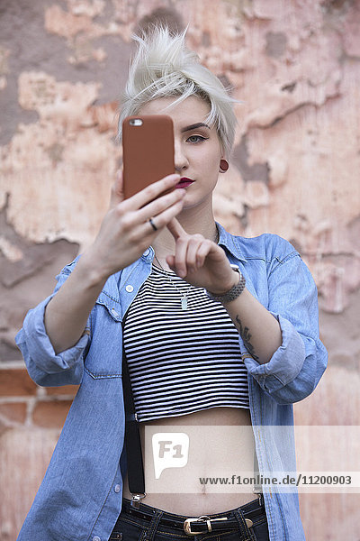 Selbstbewusste junge Frau  die sich selbst durch das Smartphone nimmt  während sie an der Wand steht.