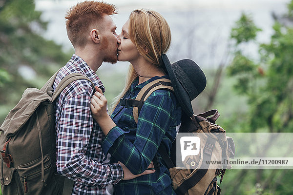 Loving couple wearing backpacks kissing while standing at forest