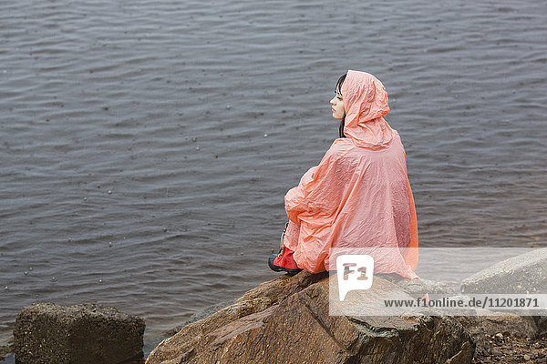 Nachdenkliche Frau im Regenmantel auf dem Felsen am Seeufer während der Regenzeit