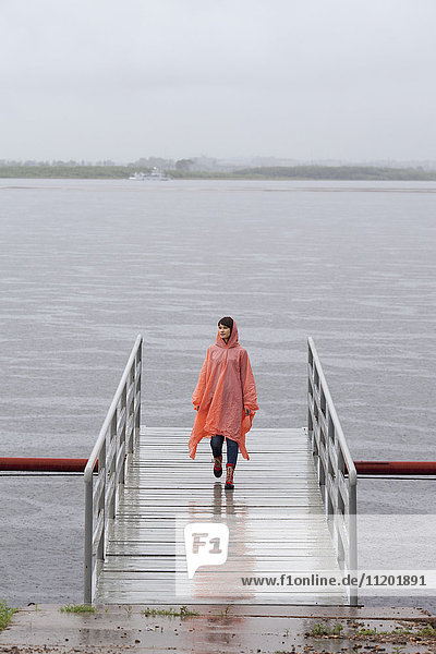 Frau im Regenmantel  die während der Regenzeit auf dem Steg läuft.