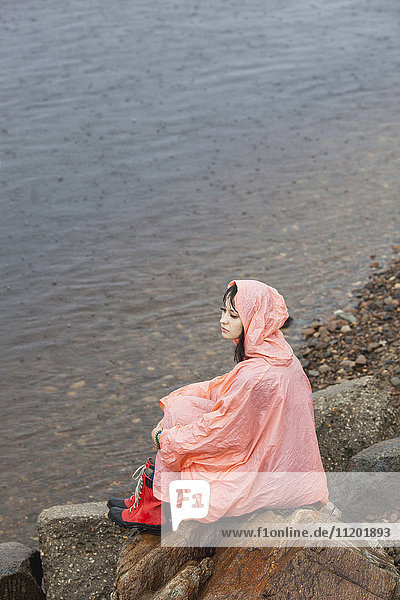 Nachdenkliche Frau im Regenmantel auf dem Felsen am Seeufer in der Regenzeit