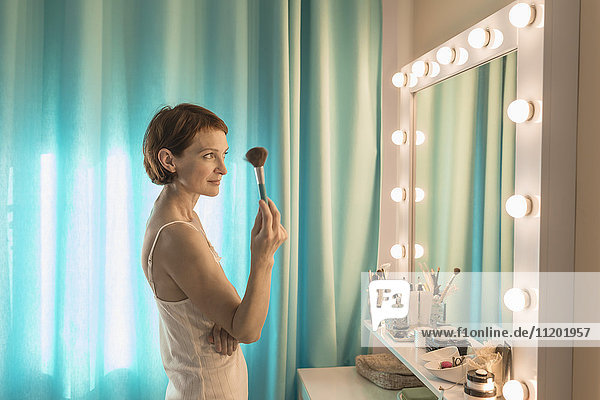 Frau hält Schminkpinsel vor dem Spiegel am Frisiertisch stehend