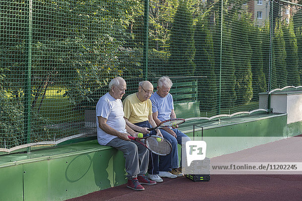 Seniorenfreunde mit Tennisschlägern  die am Hof gegen den Zaun sitzen.