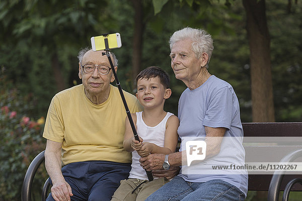 Smiling boy taking selfie by monopod with grandfathers at park bench