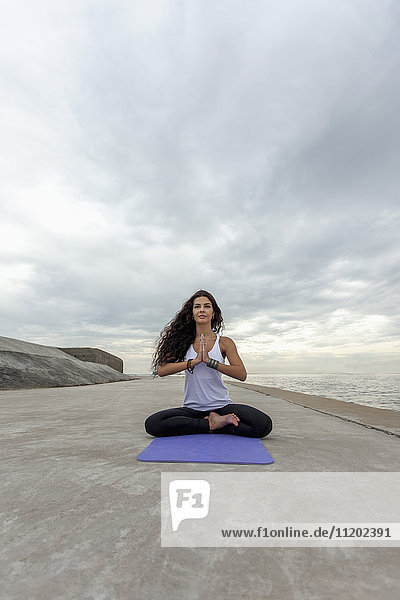 Junge Frau praktiziert Yoga in Gebetsposition am Strand gegen bewölkten Himmel