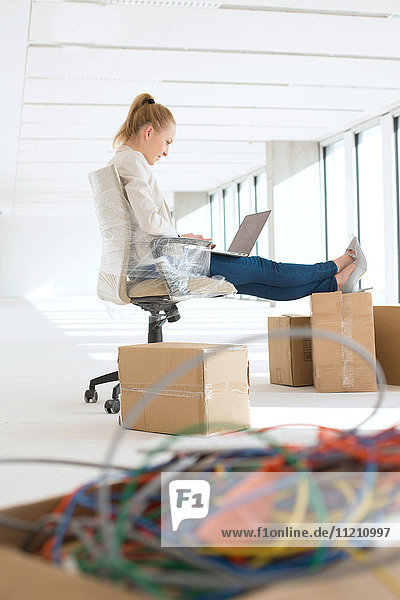 Side view of young businesswoman using laptop with feet up on cardboard box in office