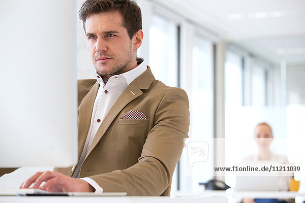Confident young businessman using computer in office with female colleague in background